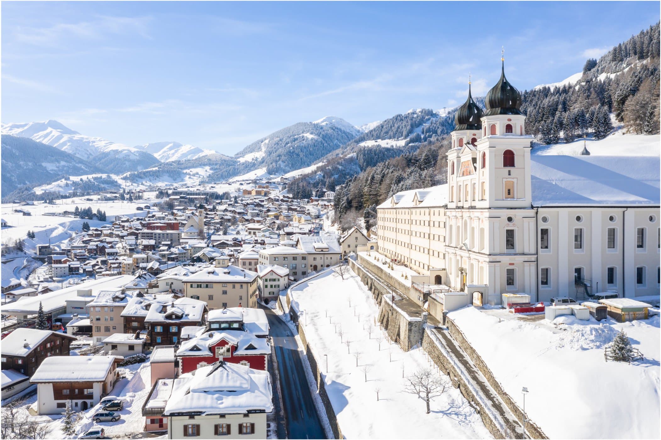 Luftaufnahme der verschneiten Stadt Disentis mit einer Kirche mit zwei Kuppeltürmen. Im Hintergrund ragen die majestätischen Alpen unter einem klaren blauen Himmel hervor und verleihen dieser malerischen Umgebung einen kulturellen Charme.