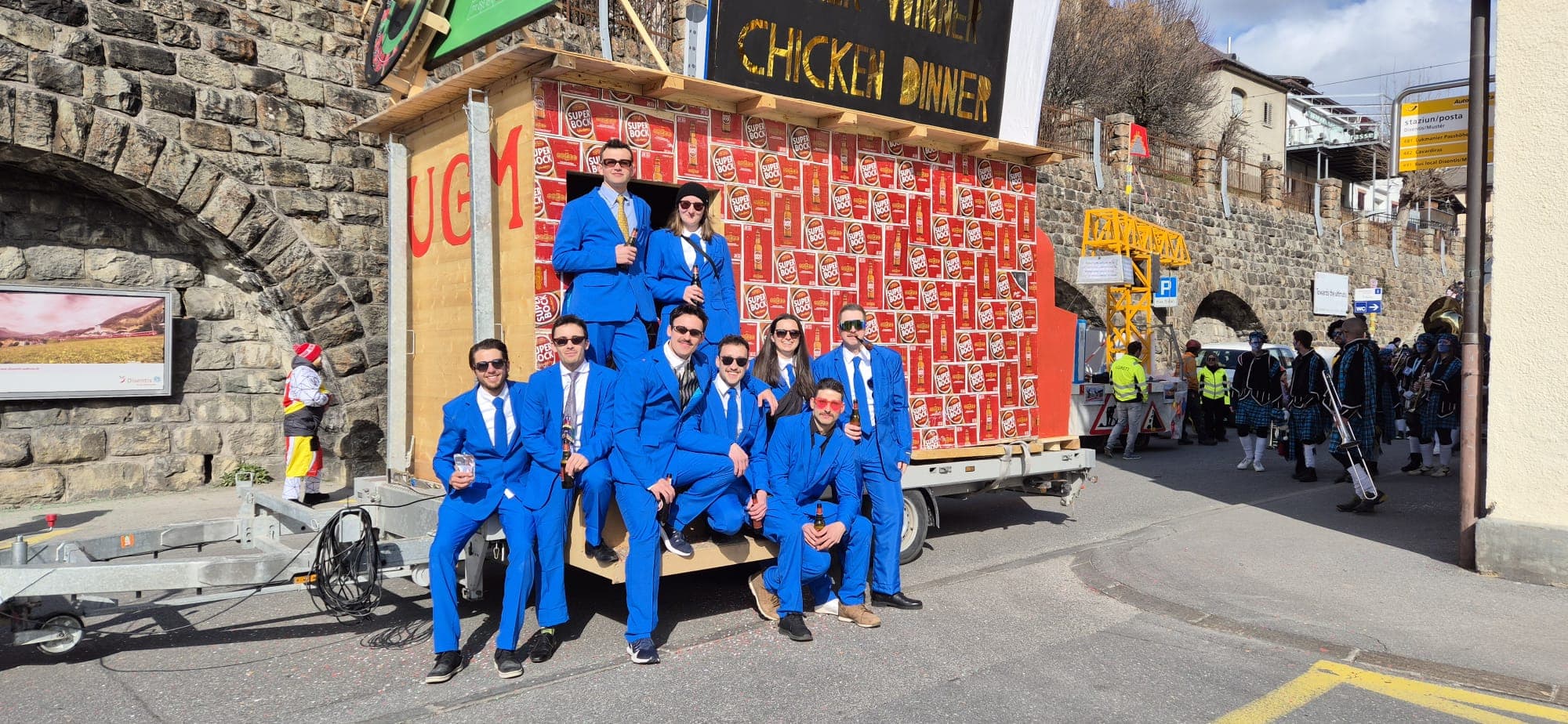 Eine Gruppe von Menschen in passenden blauen Anzügen posiert freudig vor einer mobilen Struktur, die mit roten und weißen Kisten bedeckt ist und den lebendigen Geist der Disentiser Fasnacht 2025 einfängt.